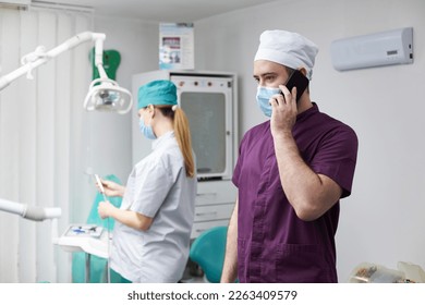 Handsome positive dentist doctor in medical mask, talks on mobile phone, standing in a modern dental office with working pregnant female dental assistant on the background - Powered by Shutterstock