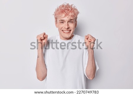 Handsome positive European man with pink hair smile broadly triumps over something clenches fists feels very glad dressed in casual basic t shirt isolated over white background. Yeah go win.