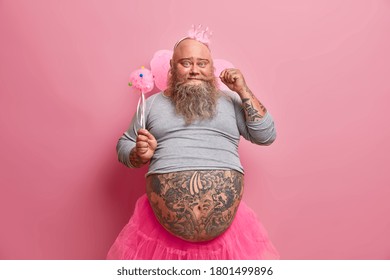 Handsome positive fat man curls mustache, has fun on party, feels like real princess, wears crown, wings, organises baby shower for her newborn daughter. Beared fatso fairy has childish mood - Powered by Shutterstock