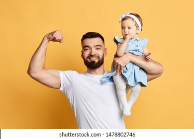 Handsome Positive Father Showing His Strength, Biceps While Holding His Cute Little Daughter. Close Up Photo. Isolated Yellow Background. Strong Bearded Man Training With His Kid In Hand