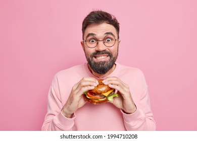 Handsome positive guy has binge eating unhealthy nutrition holds appetizing hamburger looks gladfully at camera tempting to eat junk food wears round spectacles isolated over pink background