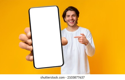 Handsome positive man showing cellphone with white blank screen and pointing on it, posing over yellow background, mockup, closeup, selective focus - Shutterstock ID 2103877637
