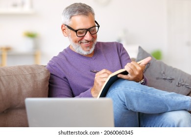 Handsome positive senior man wearing glasses working remotely while sitting on sofa with laptop computer at home, happy retired pensioner smiling  while learning studying online - Powered by Shutterstock