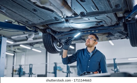 Handsome Professional Car Mechanic is Investigating Rust Under a Vehicle on a Lift in Service. Repairman is Using a LED lamp and Walks Towards. Specialist is Wearing Safety Glasses. Modern Workshop. - Powered by Shutterstock