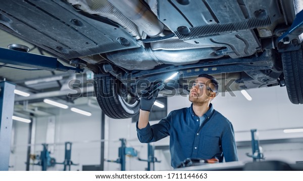 Handsome Professional Car Mechanic is\
Investigating Rust Under a Vehicle on a Lift in Service. Repairman\
is Using a LED lamp and Walks Towards. Specialist is Wearing Safety\
Glasses. Modern\
Workshop.