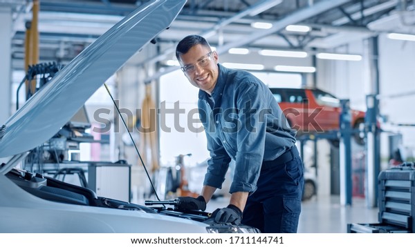 Handsome\
Professional Car Mechanic is Working on a Vehicle in a Service.\
Repairman Looks Happy While Using a Ratchet. Specialist is Wearing\
Safety Glasses. He Looks at a Camera and\
Smiles.