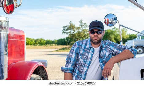 Handsome, Professional Looking Truck Driver Wearing Blue  Plaid Shirt,  Protective Sunglasses And Black American Baseball Cap Sits Next To Semi Truck. Trucker Is Next To Big Rigs, Looking At Camera.