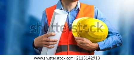 Handsome professional worker builder in special uniform holding blueprint and helmet, while standing at counstruction site indoors and checking all details according with plan.