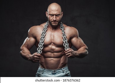 Handsome And Proud Man With Muscular Build And Bald Head Poses With Naked Torso And Steel Chains In Dark Background Looking At Camera.