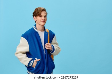 handsome red-haired smiling man student in a trendy jacket and with an orange backpack stands with his hand in his pocket on a blue background with empty space to insert advertising text