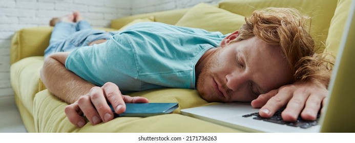 Handsome Redhead Man Napping On The Couch With Laptop And Smart Phone Laying Near Him