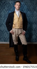 A handsome Regency gentleman wearing a gold waistcoat and black jacket and standing in a room with blue wallpaper and a wooden floor - Shutterstock ID 1960175521