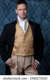 A handsome Regency gentleman wearing a gold waistcoat and black jacket and standing in a room with blue wallpaper and a wooden floor