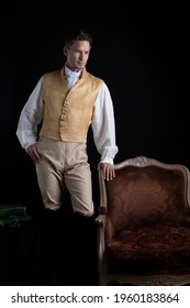 A handsome Regency gentleman wearing a linen shirt, gold waistcoat, breeches and leather boots and standing in a darkened room 