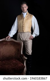 A handsome Regency gentleman wearing a linen shirt, gold waistcoat, breeches and leather boots and standing in a darkened room 