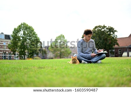 Handsome and relaxed young man reading a book while sitting in a park with his lovely cat. 	