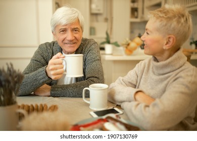 Handsome retired man with gray hair holding large mug enjoying morning tea during breakfast, sitting at dining table with his beautiful wife, talking, having nice conversation. People and family - Powered by Shutterstock