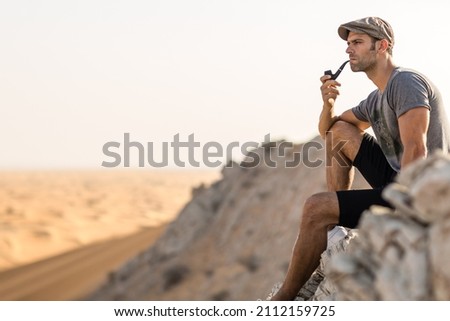 Handsome retro man sitting alone smoking a pipe on top of a rocky mountain overlooking the vast desert view.
