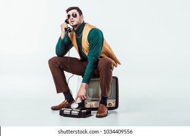handsome retro styled man sitting on vintage television and talking by stationary telephone 