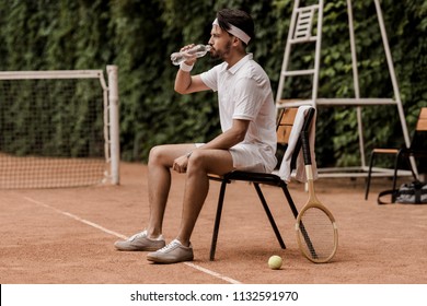 Handsome Retro Styled Tennis Player Sitting On Chair And Drinking Water At Tennis Court