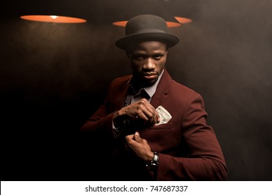 handsome rich african american man in hat holding dollar banknotes in smoky room with lamps