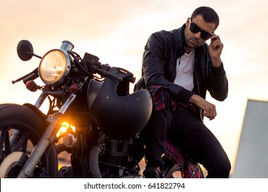 Handsome rider guy with beard and mustache in black biker jacket take off sunglasses on classic style cafe racer motorcycle at sunset. Bike custom made in vintage garage. Brutal fun urban lifestyle. - Powered by Shutterstock