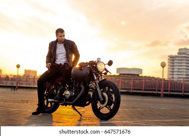 Handsome Rider Guy With Beard And Mustache In Black Leather Biker Jacket Sit On Classic Style Cafe Racer Motorcycle At Sunset Time. Bike Custom Made In Vintage Garage. Brutal Fun Urban Lifestyle.