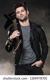 handsome rocker holding electric guitar and looking at camera on dark smoky background
