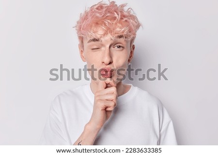 Handsome romantic man keeps lips folded hand on chin winks eye wants to kiss girfriend flirts with someone has pink wavy hair dressed in white t shirt poses indoor. Facial expressions concept