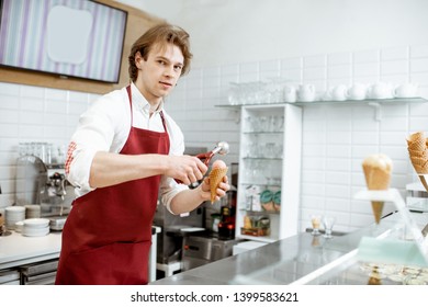 Handsome Salesman In Red Apron Making Ice Cream In Waffle Cone For Selling In The Modern Pastry Shop