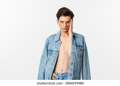 Handsome and sassy gay man wearing denim jacket on bare torso, touching his face and looking confident at camera, standing over white background - Powered by Shutterstock