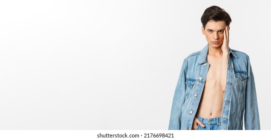 Handsome and sassy gay man wearing denim jacket on bare torso, touching his face and looking confident at camera, standing over white background - Powered by Shutterstock