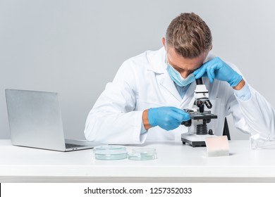 handsome scientist in medical mask and medical gloves looking at microscope isolated on white