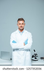 handsome scientist in white coat and latex gloves standing with crossed arms and looking at camera isolated on white