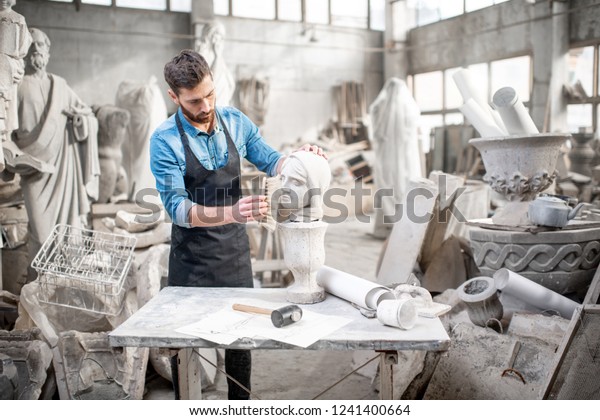 Handsome sculptor brushing stone head\
sculpture on the table in the atmospheric\
studio