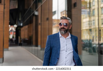 A handsome senior business man in glasses and a suit is walking through the city street near the office building. - Shutterstock ID 2216002311