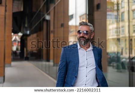 A handsome senior business man in glasses and a suit is walking through the city street near the office building.