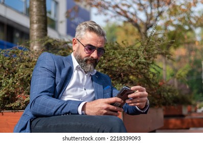 A Handsome Senior Business Man Using Mobile Phone App Texting While Sitting On The Park Bench.