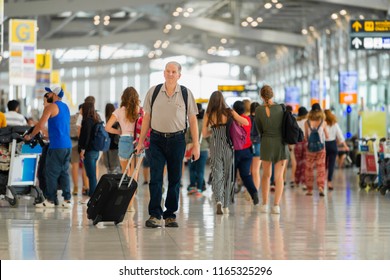 Handsome Senior Man With Backpack Walking With A Luggage At Airport Terminal And Airport Terminal Blurred Crowd Of Travelling People On The Background.  