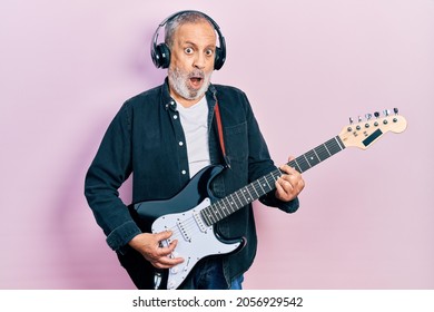 Handsome senior man with beard playing electric guitar in shock face, looking skeptical and sarcastic, surprised with open mouth  - Powered by Shutterstock