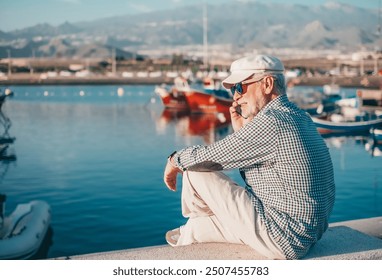 Handsome senior man in casual shirt and hat talking on phone while sitting at the port at sea. Relaxed elderly bearded male wearing sunglasses enjoying freedom, travel or retirement - Powered by Shutterstock