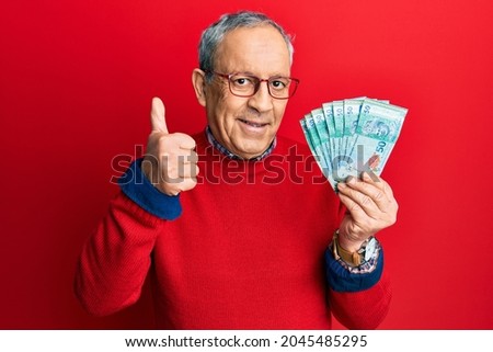 Handsome senior man with grey hair holding 50 malaysia ringgit banknotes smiling happy and positive, thumb up doing excellent and approval sign 