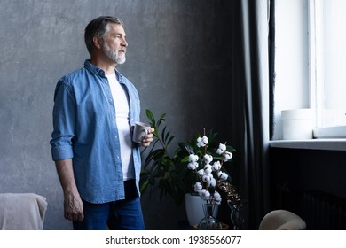 Handsome senior man is holding a cup, looking away and smiling while standing in apartment. - Powered by Shutterstock