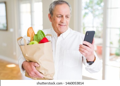 Handsome senior man holding paper bag full of fresh groceries and looking at smartphone smiling - Powered by Shutterstock