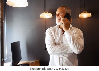 Handsome senior man talking on phone and smilling. CEO works in modern loft style office using devices and gadgets during job processing. Success, corporate teamwork, age and experience.