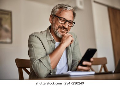 Handsome senior man wearing glasses using smartphone while sitting at his cozy workplace with laptop at home. - Powered by Shutterstock