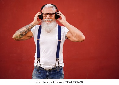 Handsome Senior Man With White Beard, Mustache And With Tattoos Listening To Music With Headphones. Concept Of Elderly People Lifestyle And Hobby. Real People. Red Background. Copy Space.
