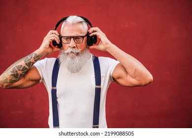 Handsome Senior Man With White Beard, Mustache And With Tattoos Listening To Music With Headphones. Concept Of Elderly People Lifestyle And Hobby. Real People. Red Background. Copy Space.
