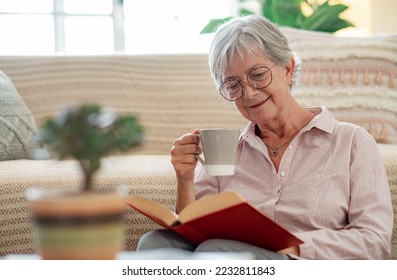 Handsome senior woman sitting on the floor at home reading a book while enjoying a cup of tea. Smiling mature lady relaxed in retirement - Powered by Shutterstock