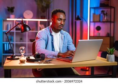 Handsome serious african man in casual clothes sitting at table and typing on wireless laptop. Young guy doing remote work or studying online during evening time at home. Freelance concept.